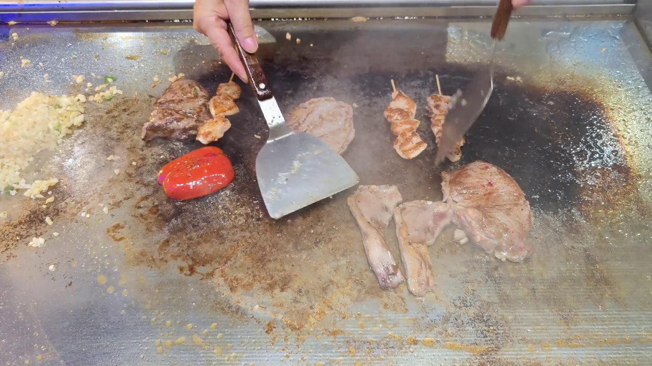 Tenderloin, steak, chicken skewers, peppers and lamb chops are topped with sliced ​​garlic and spring onions by an unrecognizable chef and then fried on a teriyaki grill plate in a restaurant视频素材画面3