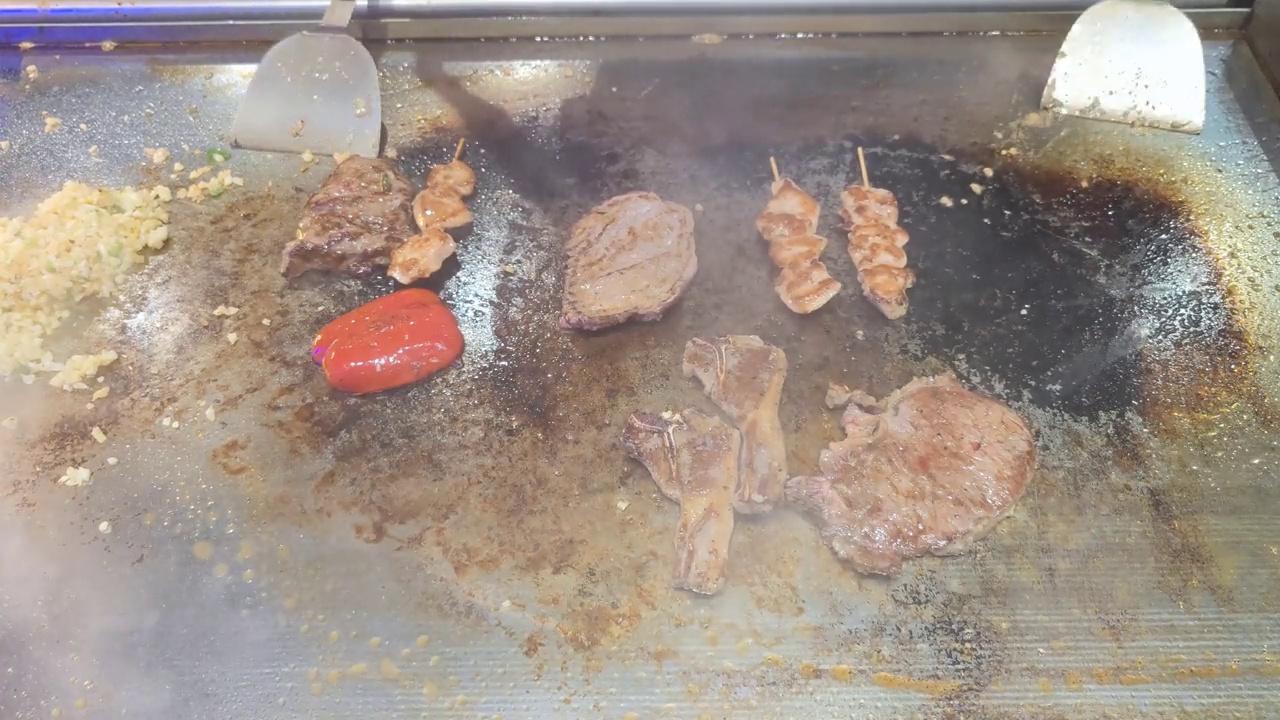 Tenderloin, steak, chicken skewers, peppers and lamb chops are topped with sliced ​​garlic and spring onions by an unrecognizable chef and then fried on a teriyaki grill plate in a restaurant视频素材画面4