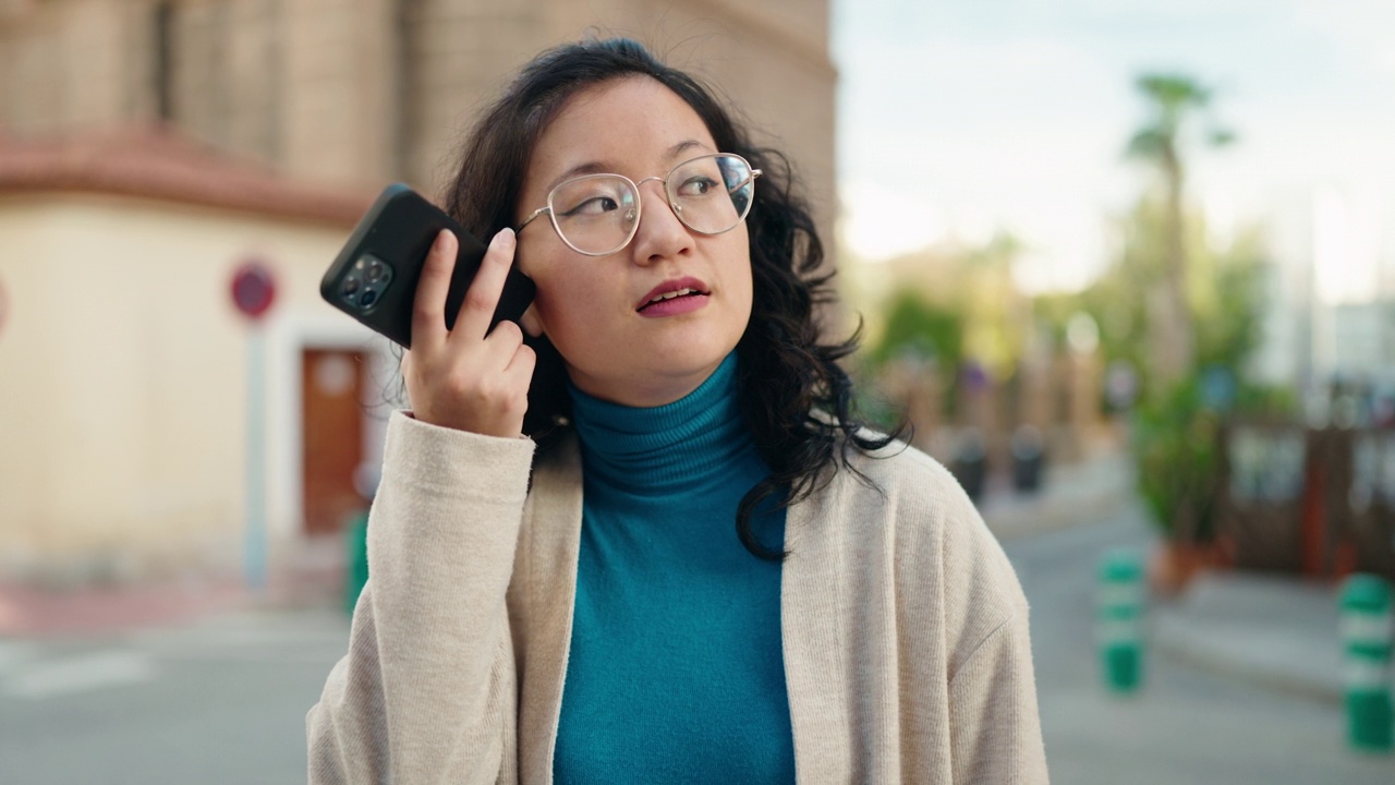 年轻的中国女子微笑着自信地在街上通过智能手机听音频信息视频素材