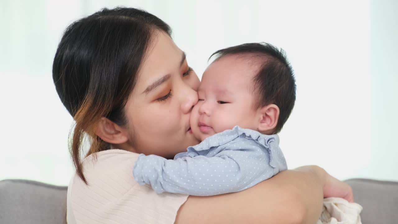 爱温柔，年轻的亚洲母亲抱着可爱可爱的女婴女儿拥抱，玩耍和亲吻小孩。快乐的亚洲母亲抱着婴儿在家里客厅视频素材