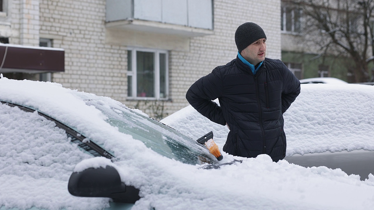 那个男人一边清理汽车上的积雪，一边感到背部疼痛。视频素材