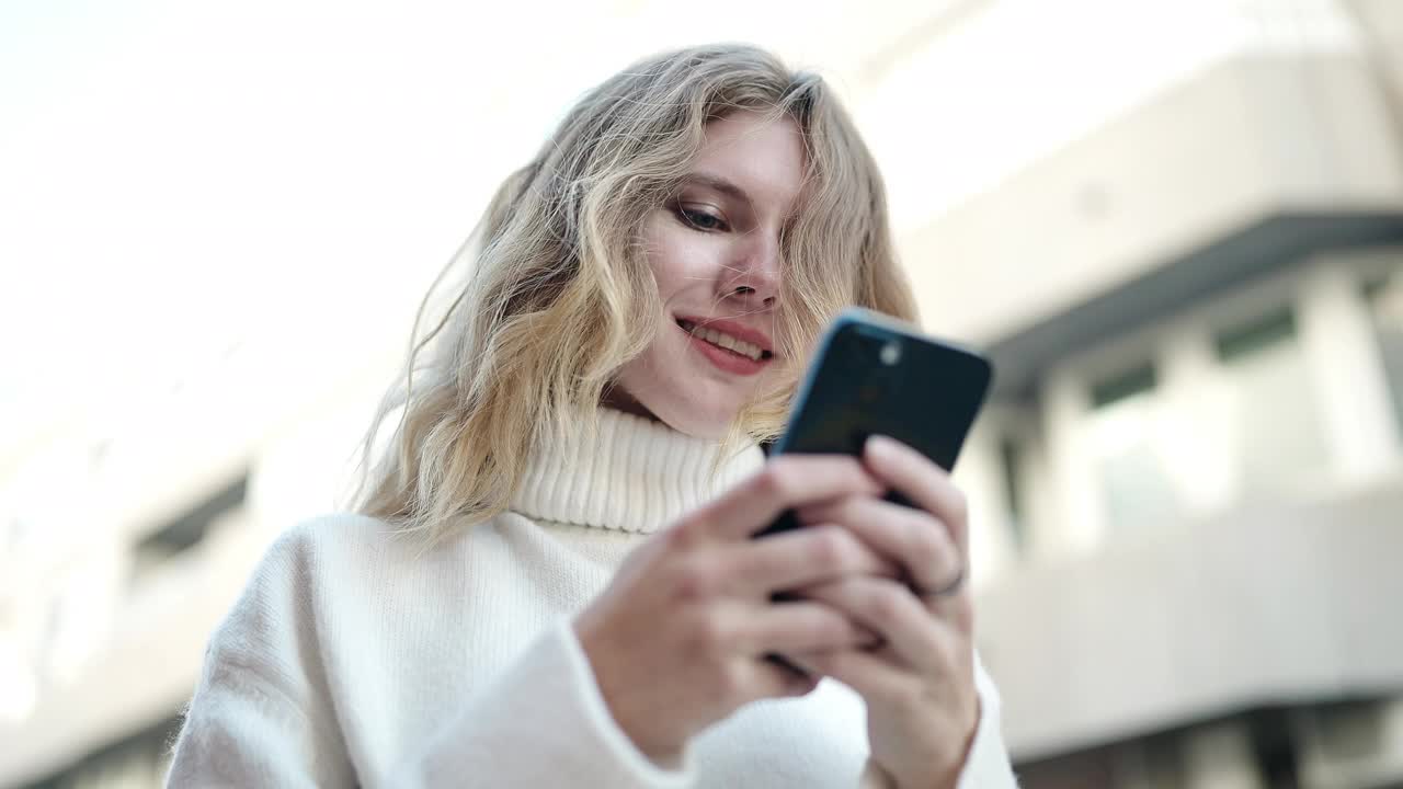 年轻的金发女子在街上自信地微笑着使用智能手机视频素材