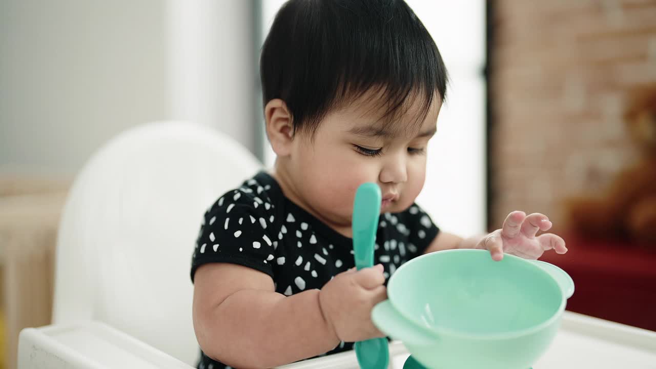 可爱的西班牙婴儿坐在家里抱着勺子的高脚椅上视频素材