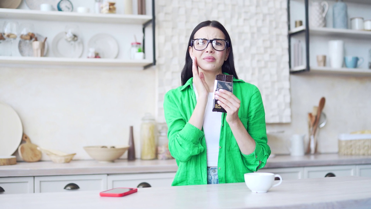 年轻的黑发女人在绿色衬衫吃巧克力和有牙痛在光明的家庭厨房。悲伤的女孩感到牙齿疼痛，因为龋齿视频素材