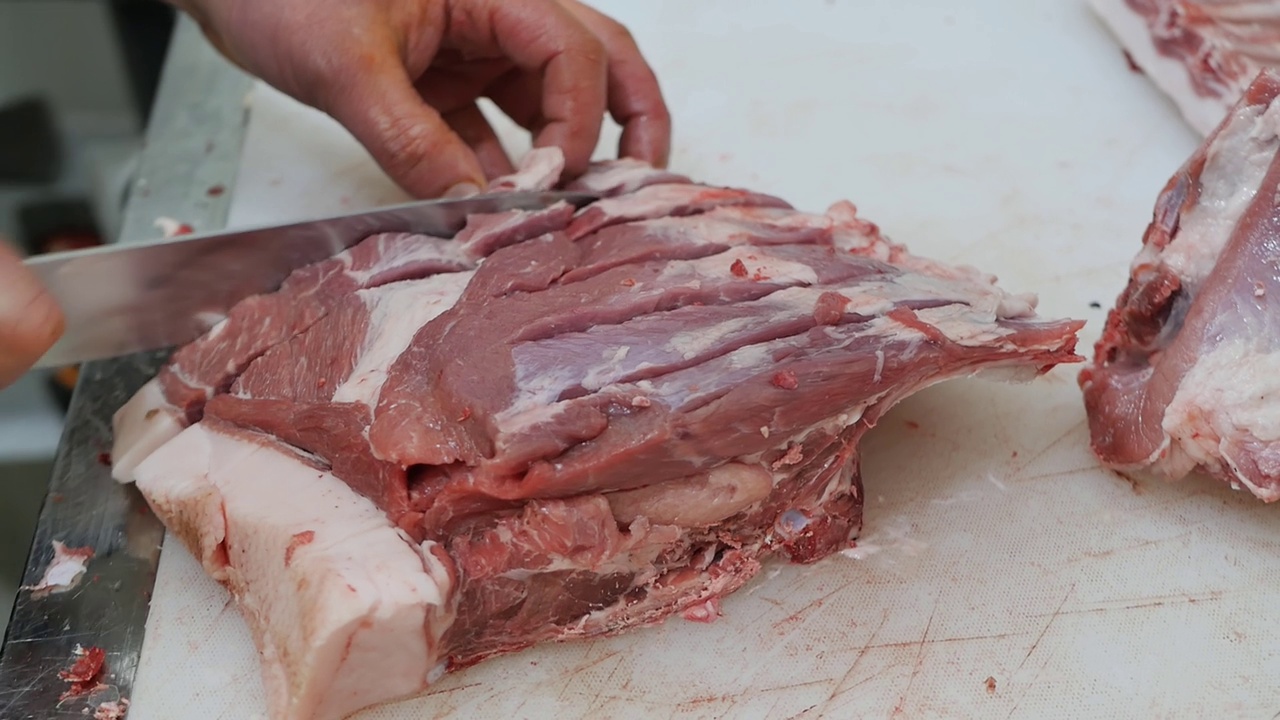 在熟食店的肉铺里，屠夫用一把锋利的大刀在砧板上切生肉的过程特写。视频素材