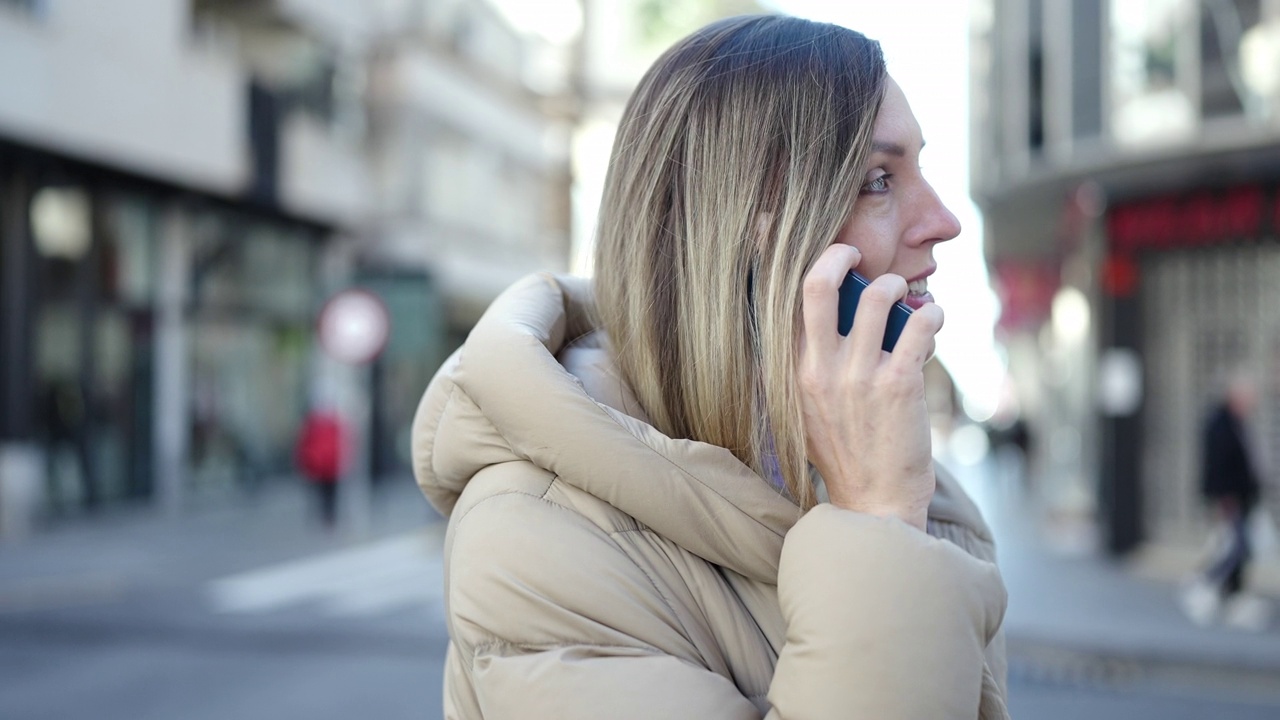 年轻的金发女人微笑着自信地在街上用智能手机说话视频素材