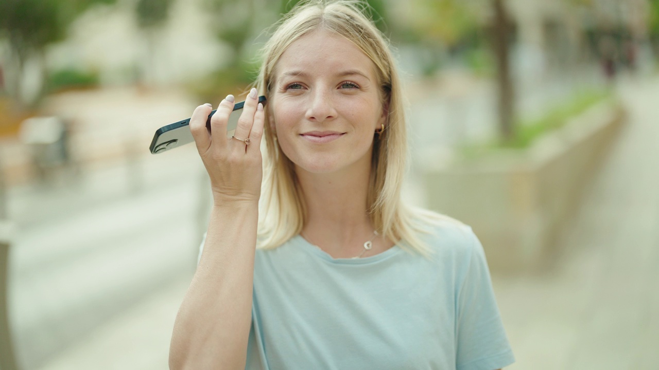 年轻的金发女人微笑自信地听着智能手机在街上的音频信息视频素材