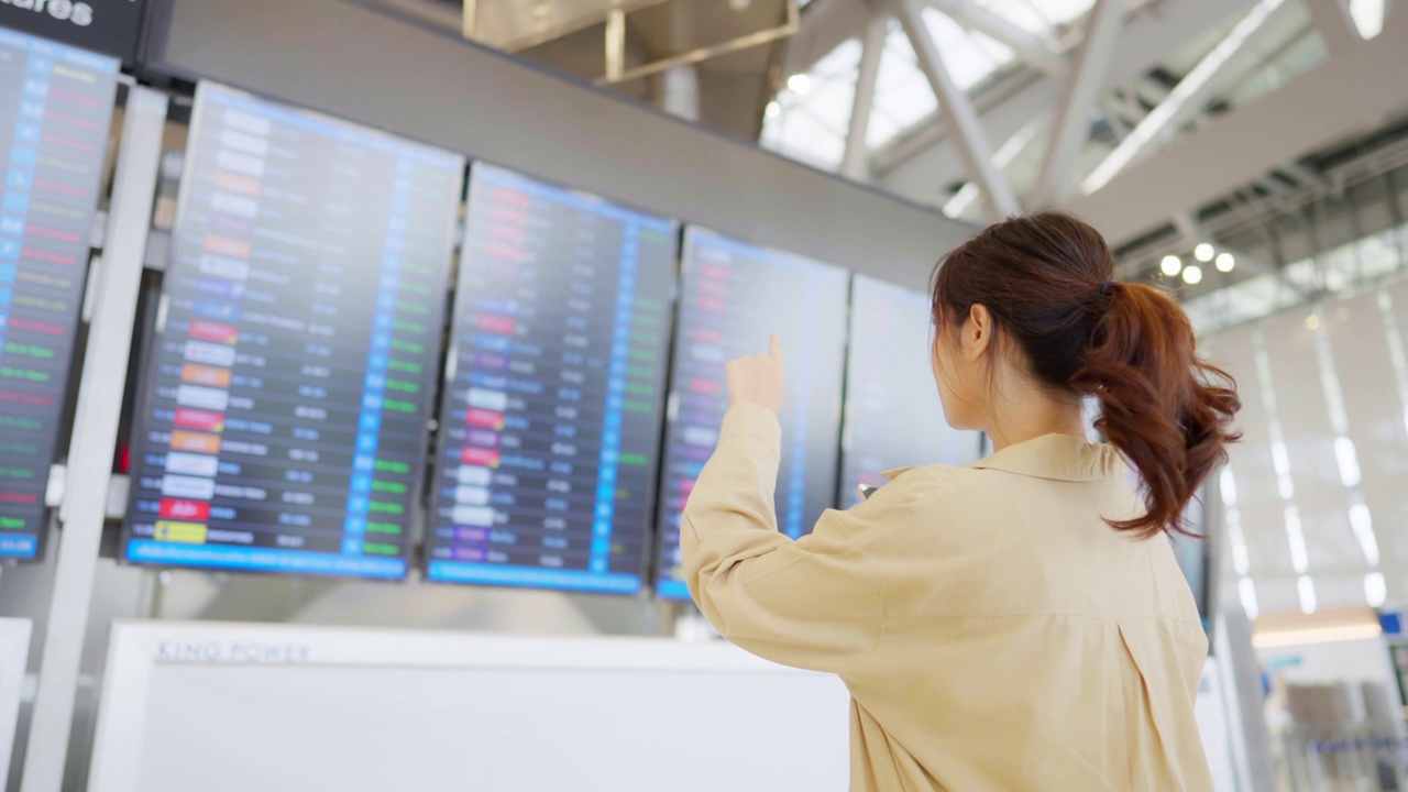 年轻的亚洲女子拿着护照和登机牌在国际机场看航班信息板，检查她的航班视频素材