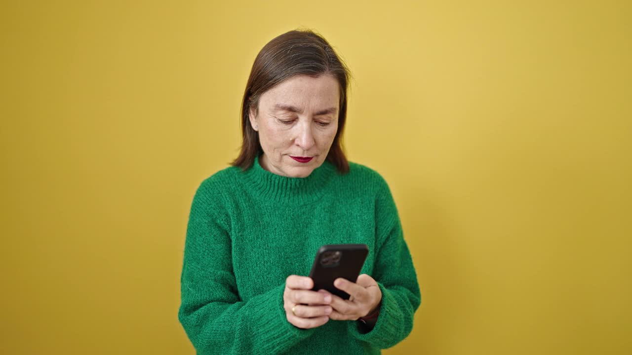 成熟的西班牙女子与灰色的头发使用智能手机微笑孤立的黄色背景视频素材