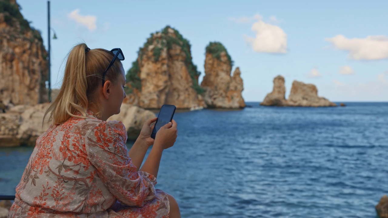 金发游客在海边度假时使用智能手机的侧面视图。女人在Scopello - Spiaggia dei Faraglioni视频素材