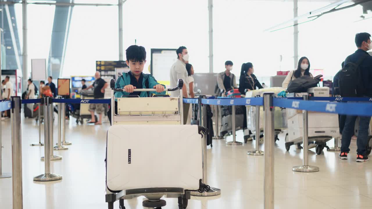 旅行是一种永无止境的学习经历。穿着休闲夹克的亚洲男孩推着行李车在机场柜台排队办理登机手续。在学校假期和家人一起旅行。视频素材