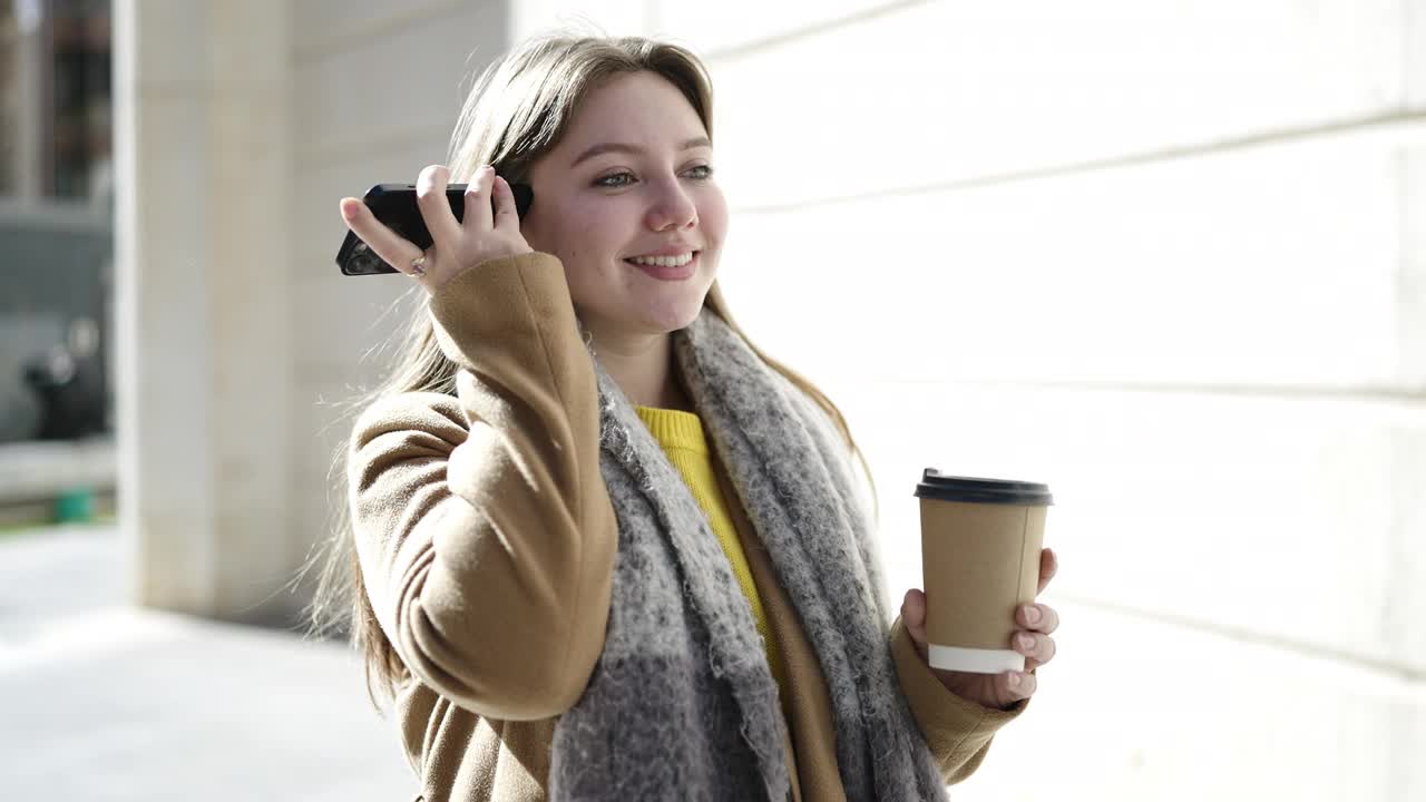 年轻的金发女子在街上用智能手机听着语音信息喝着咖啡视频素材