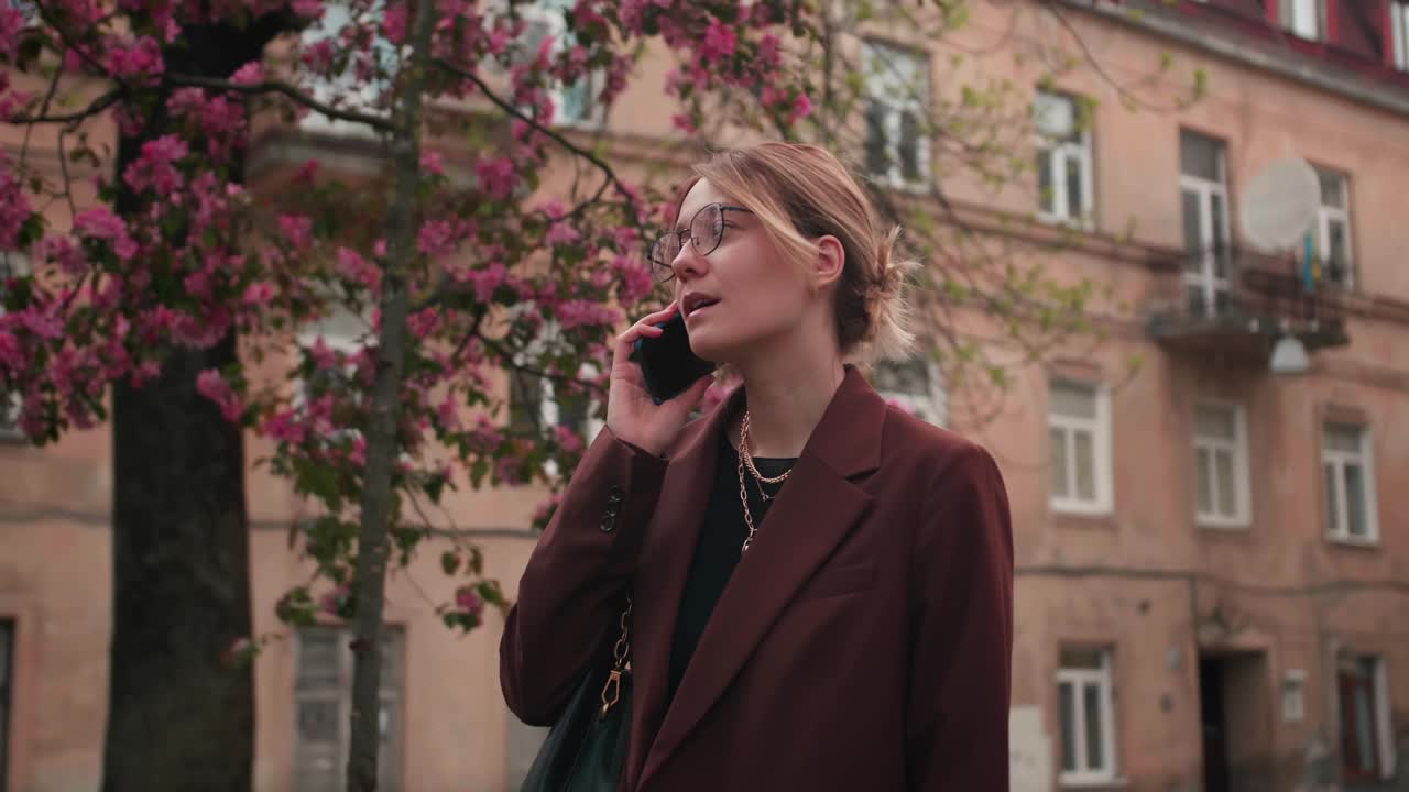 下图年轻微笑的白人女子戴着眼镜在春天公园户外休息讲手机。人们对城市生活方式的观念。视频素材