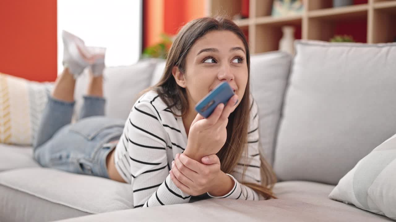 年轻漂亮的西班牙女子躺在家里的沙发上用智能手机发语音信息视频素材