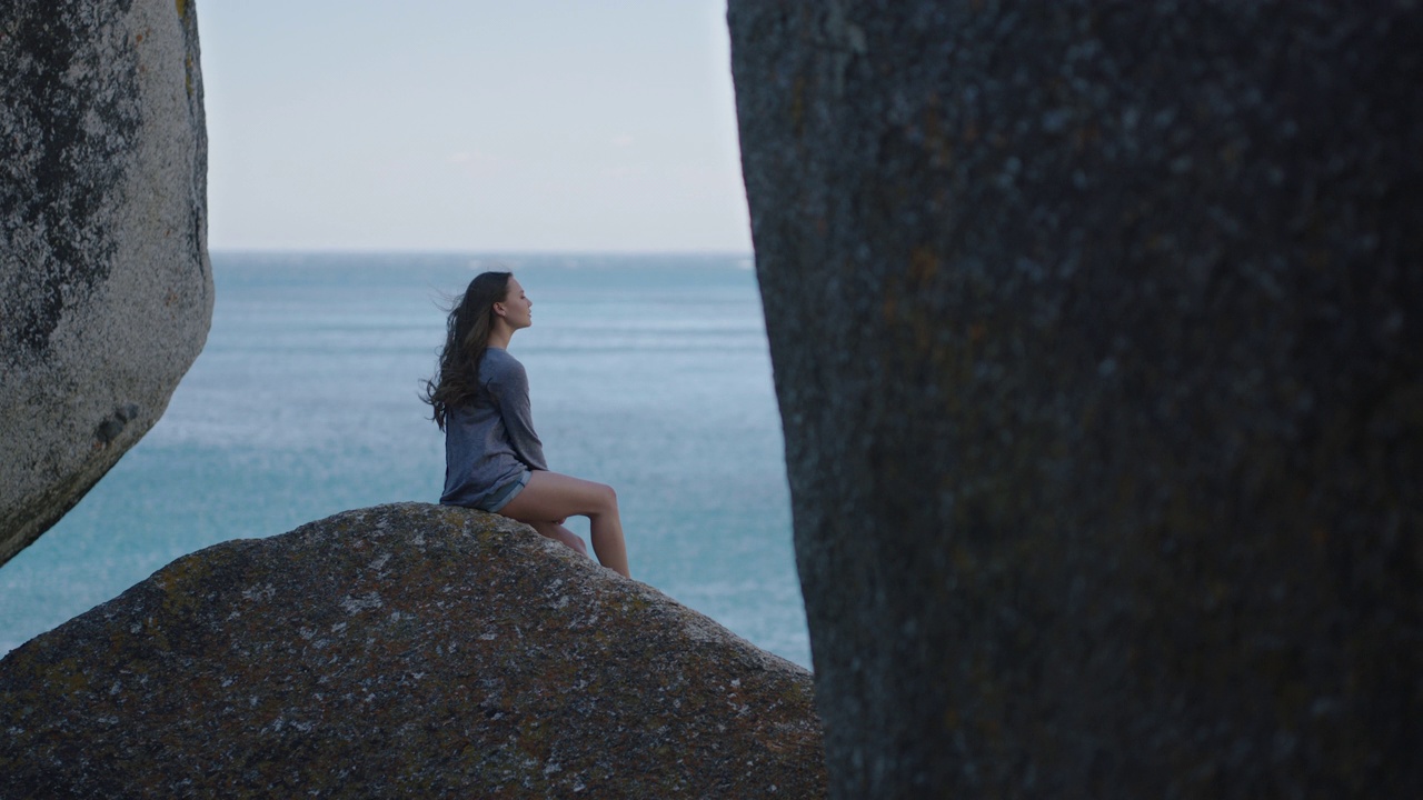 慢动作的年轻美女游客看着大海，享受宁静的海景，海风吹着头发，坐在岩石上放松假期视频素材