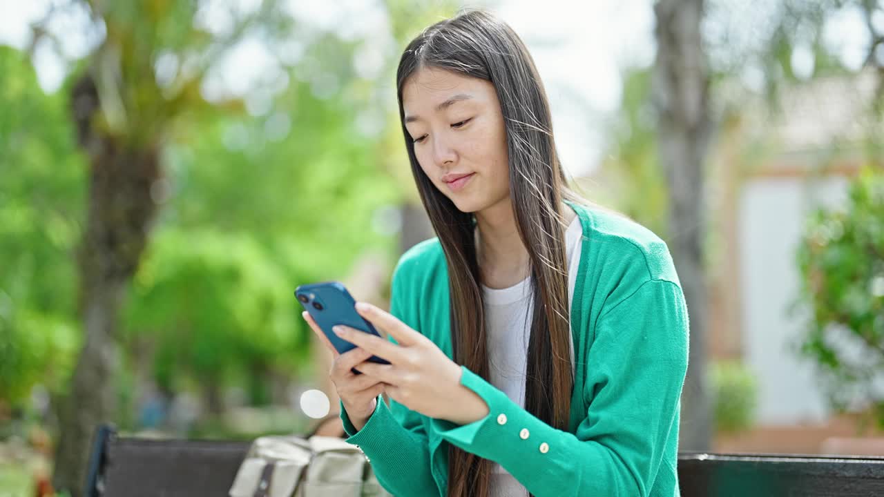 年轻的中国女子坐在公园的长椅上玩智能手机视频素材