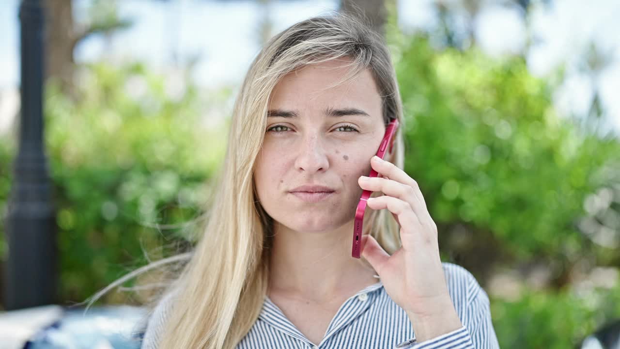 年轻的金发女子微笑着自信地在公园用智能手机聊天视频素材