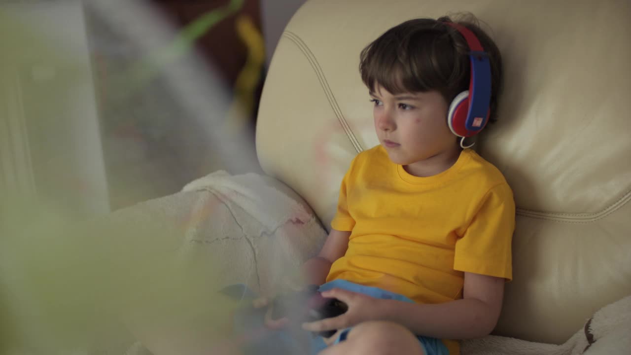 小男孩在炎热的天气里在风扇旁边玩游戏机。炎炎夏日，孩子在家用手柄玩电脑游戏。孩子坐在家里客厅的沙发上，天气炎热。视频素材