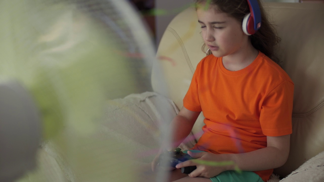 小女孩在炎热的天气里在风扇附近玩游戏机。炎炎夏日，孩子在家用手柄玩电脑游戏。孩子坐在家里客厅的沙发上，天气炎热。视频素材