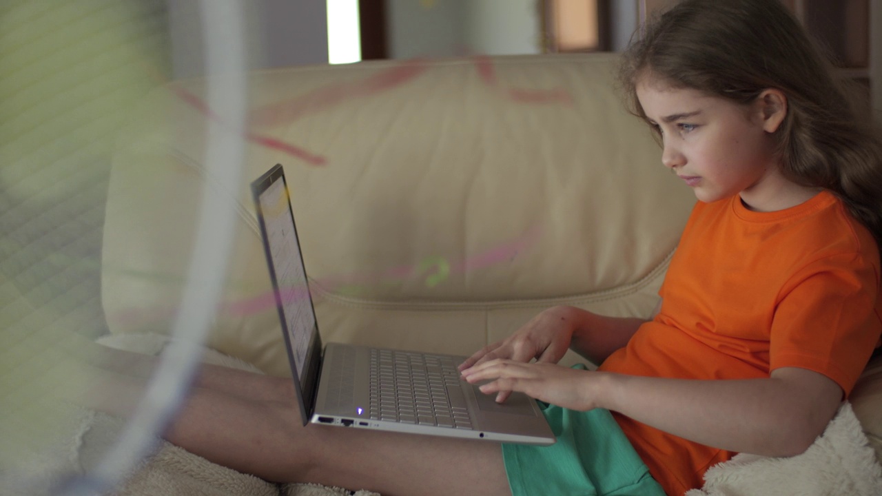 孩子坐在家里的沙发上使用笔记本电脑浏览社交媒体，在家上学。酷暑的孩子享受着风扇吹来的清凉微风。大热天在家里的客厅里视频素材