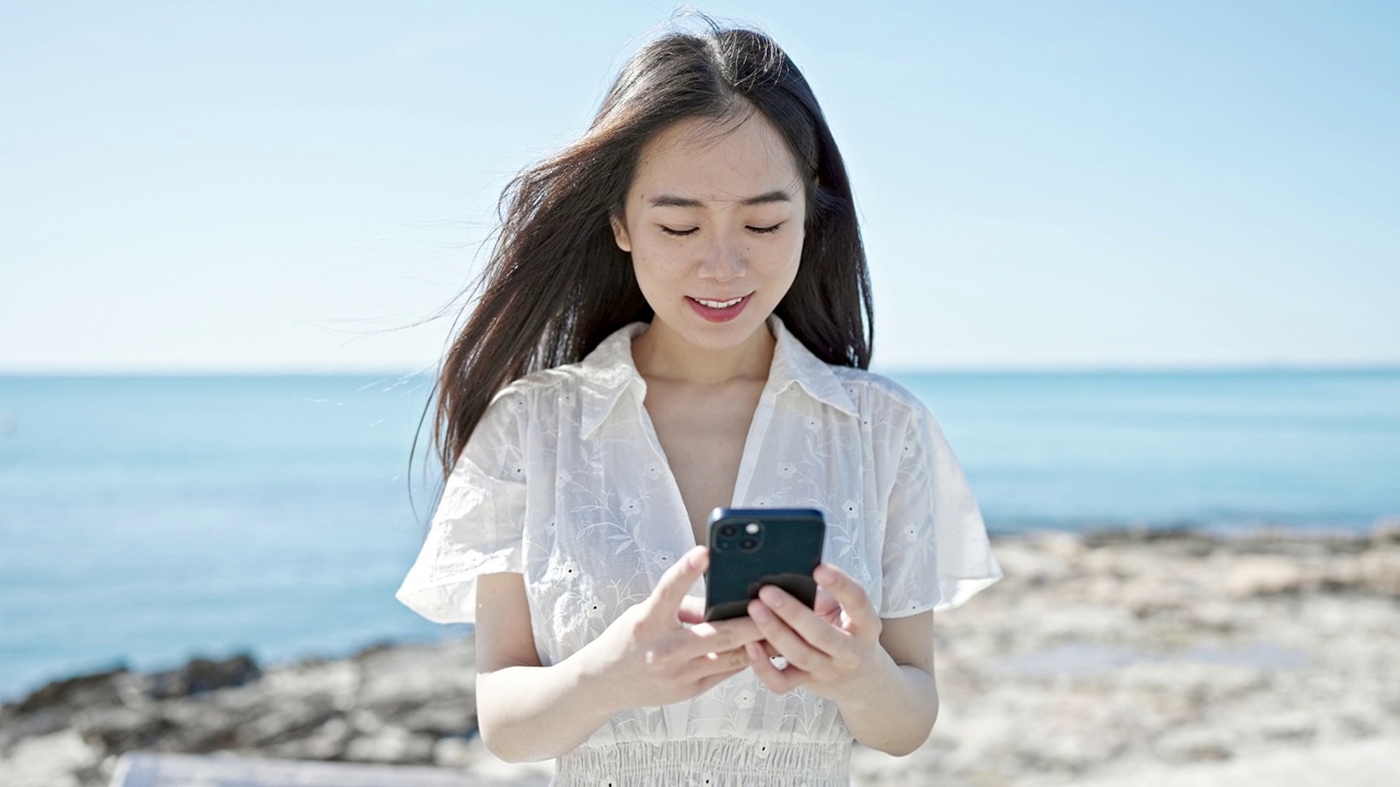 年轻的中国女子在海边用智能手机微笑视频素材