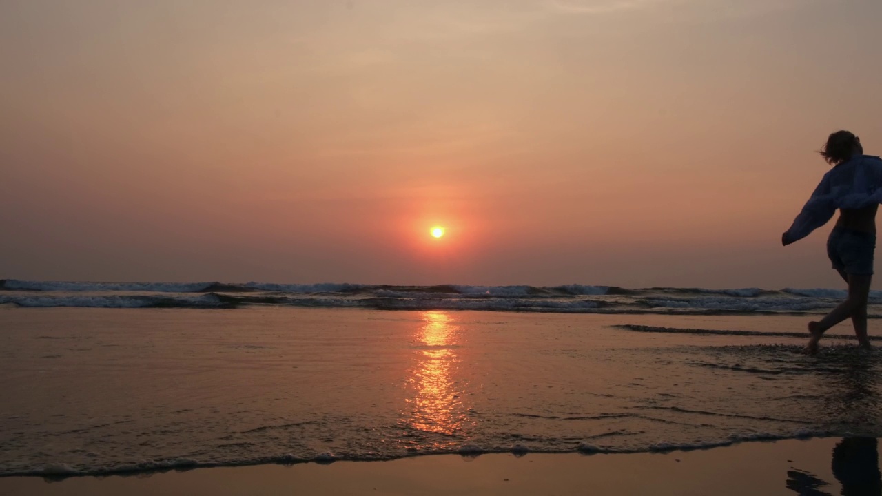 在夕阳的背景下，一个女人在海上行走的剪影。日落时分，一名女游客沿着海滩散步。视频素材