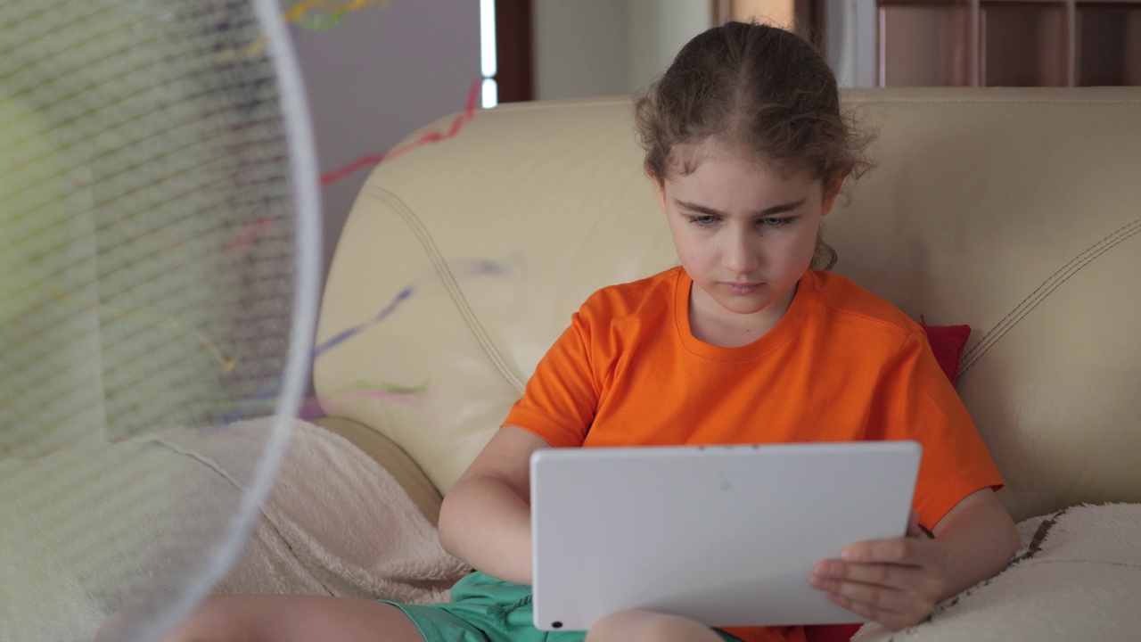 女孩在炎热的夏天坐在沙发上，坐在风扇旁边，用平板电脑浏览社交媒体。孩子在炎热的天气里在风扇旁玩平板电脑。酷暑时电风扇吹出的凉风。视频素材