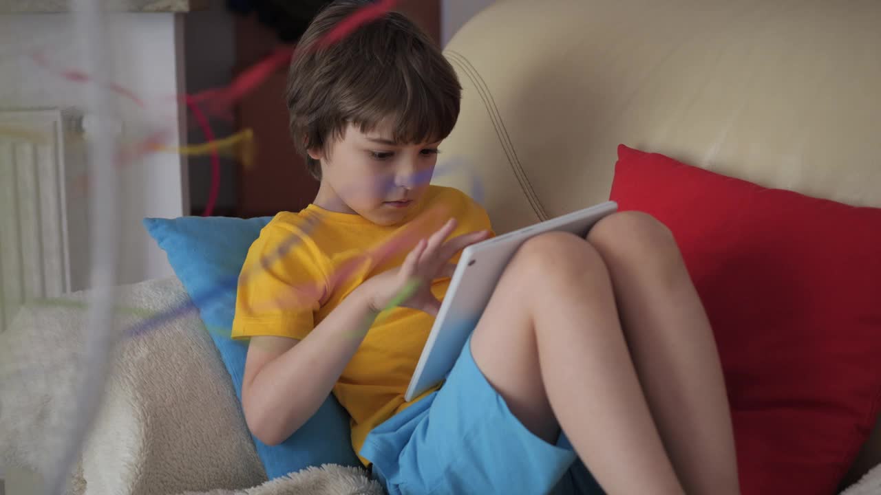 在炎热的夏天，男孩坐在沙发上，坐在风扇旁边，用平板电脑浏览社交媒体。孩子在炎热的天气里在风扇旁玩平板电脑。酷暑时电风扇吹出的凉风。视频素材