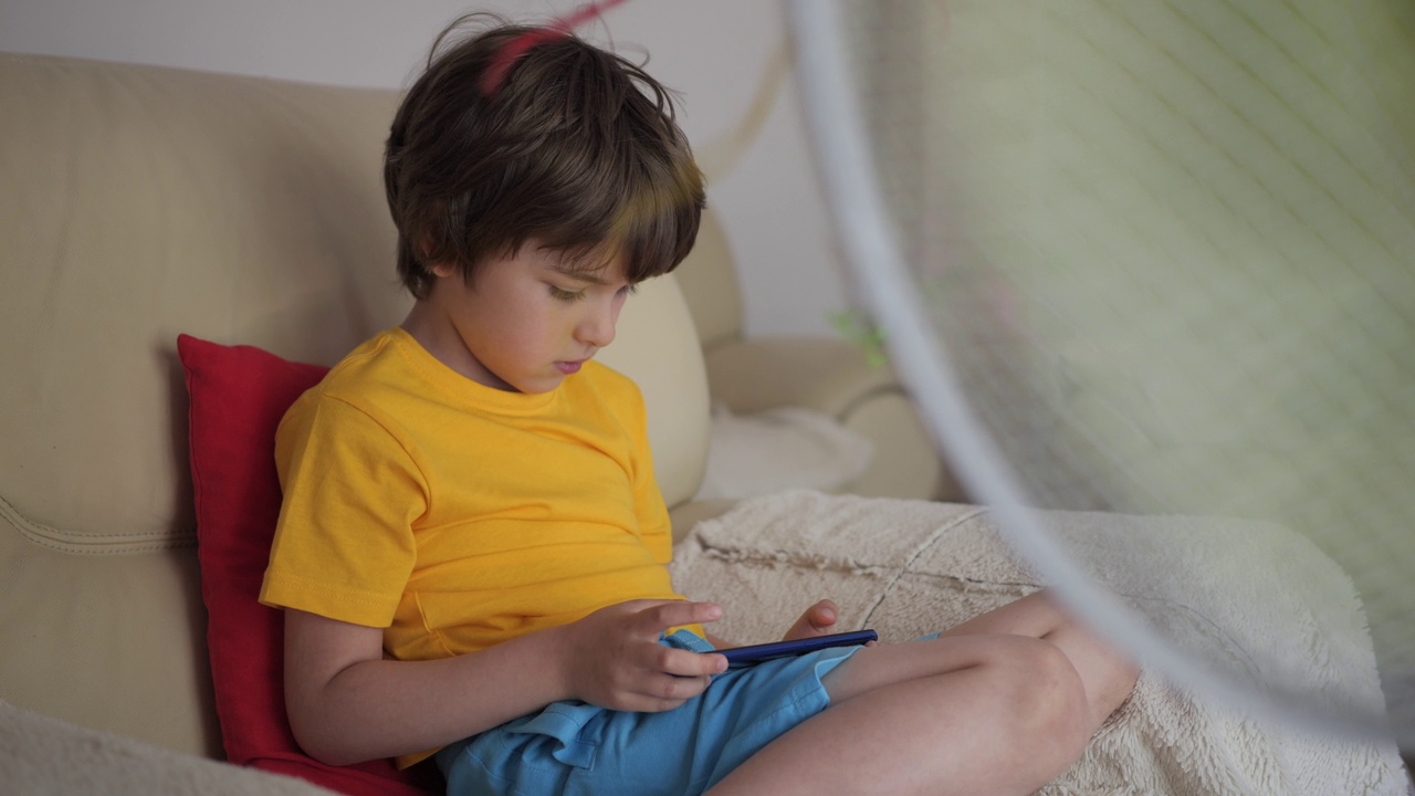 在炎热的夏天，男孩坐在沙发上靠近风扇玩智能手机游戏。在炎热的天气里，小孩在风扇旁边玩手机游戏。酷暑时电风扇吹出的凉风视频素材