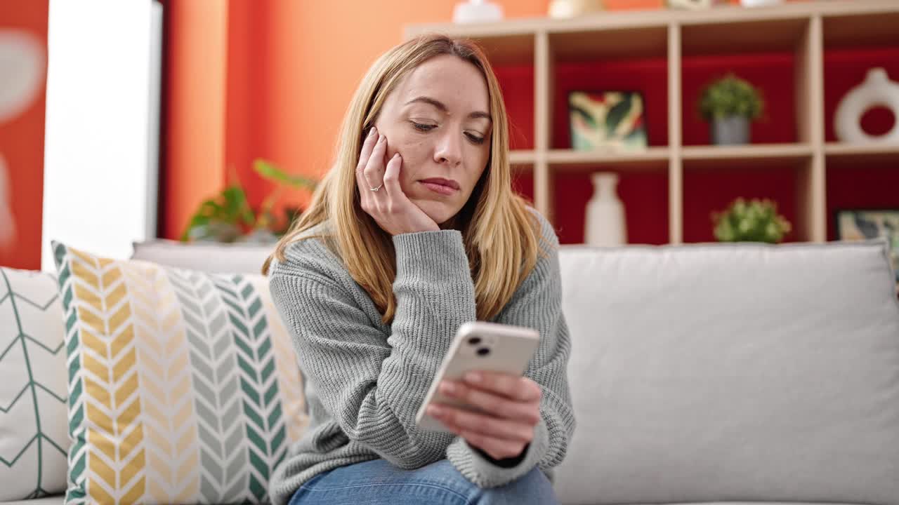 年轻的金发女子坐在沙发上玩智能手机，看起来很沮丧视频素材