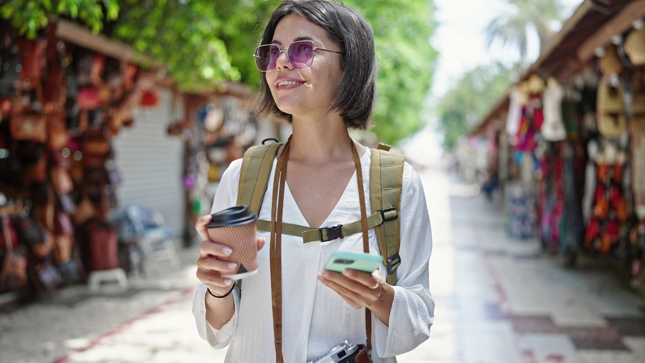 年轻美丽的西班牙女游客微笑着自信地用智能手机在街头市场喝咖啡视频素材