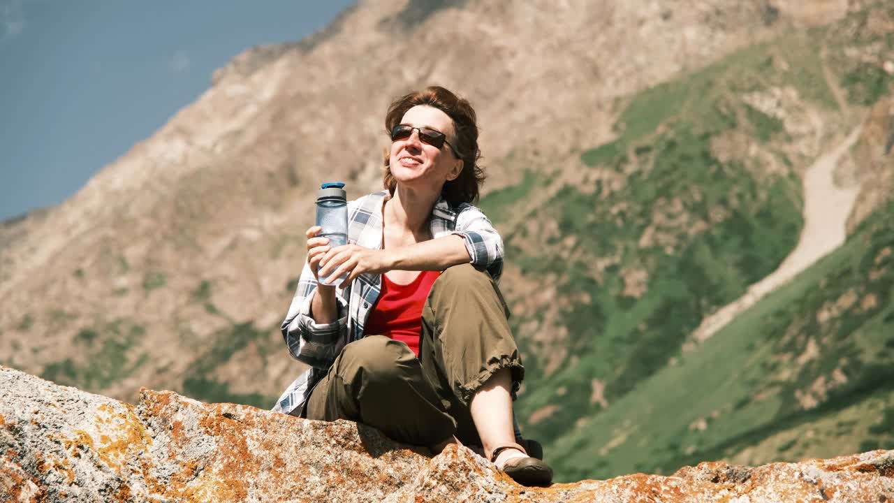 快乐的46岁女游客坐在高山上的岩石上喝水视频素材