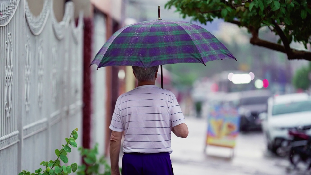 老人的背影在雨中打着伞走着。上了年纪的人在雨中漫步在人行道上视频素材