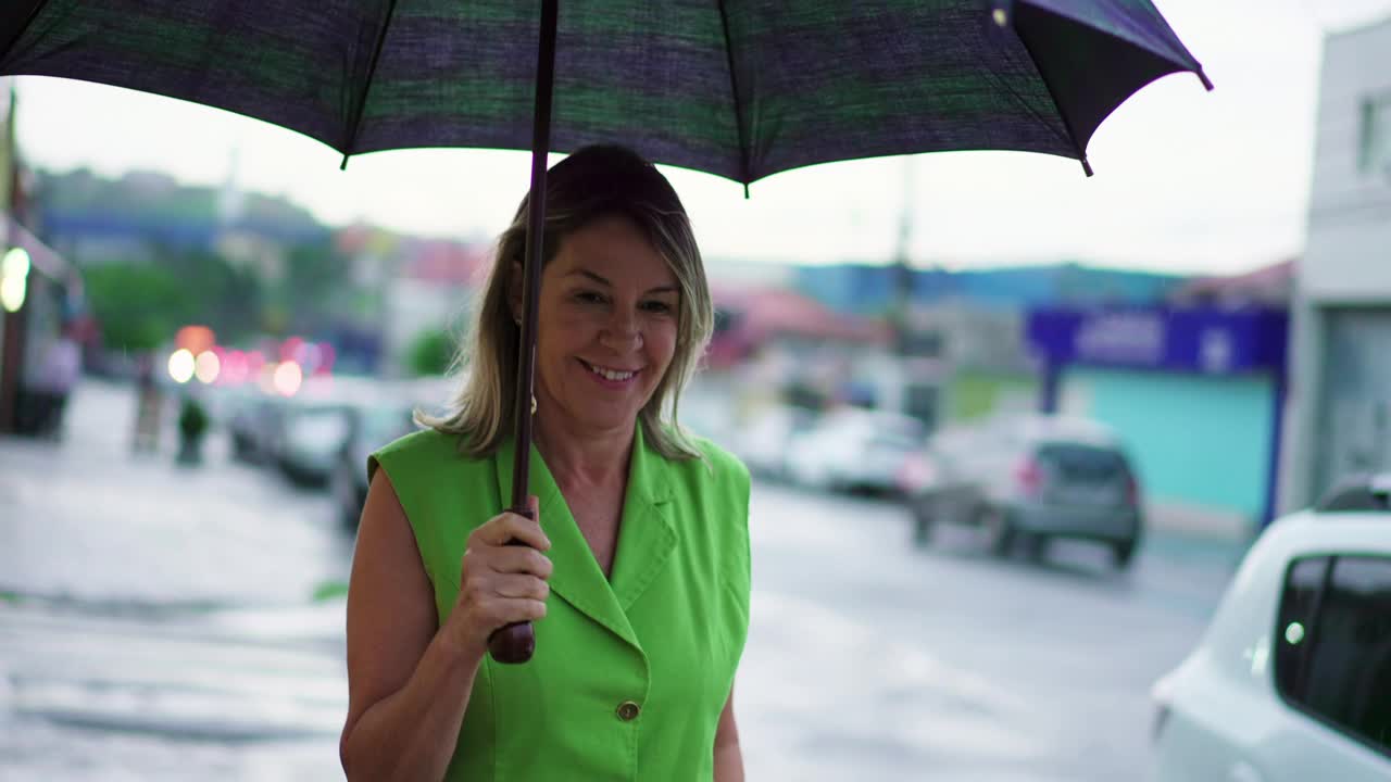 快乐的中年妇女打着伞走在雨中的城市街道人行道上视频素材