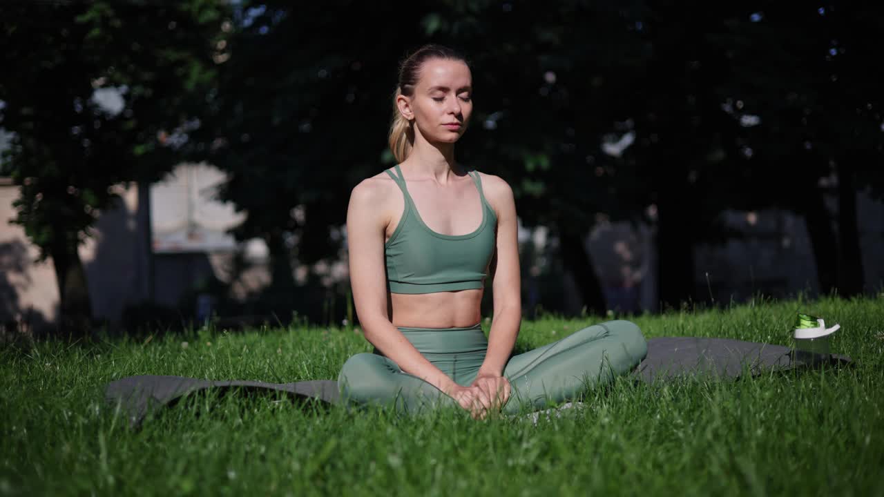 运动的美丽苗条的女人坐在瑜伽垫上莲花的姿势，在城市公园外举起双手。女运动员在阳光明媚的日子里在户外练习瑜伽。莲花体式，冥想观。视频素材