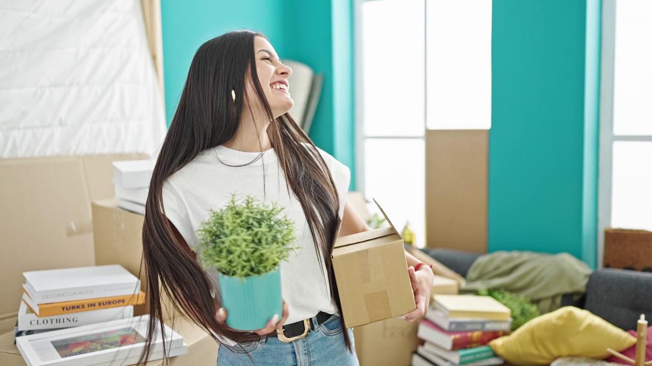 年轻美丽的西班牙女人微笑自信地拿着包裹和植物在新家视频素材