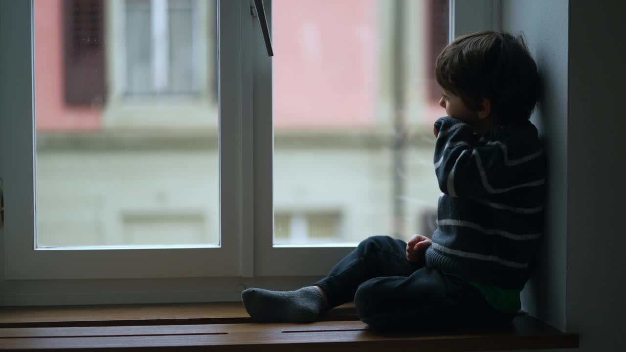 孩子坐在公寓窗边，凝视着家里细雨蒙蒙的景色。在寒冷的日子里，小男孩穿着毛衣凝视着风景视频素材