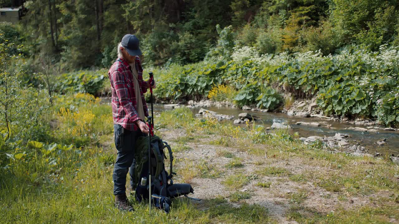 一个金发碧眼、留着胡子、穿着红色登山衫的游客在森林河附近的草坪上散步，他打开了背包视频素材