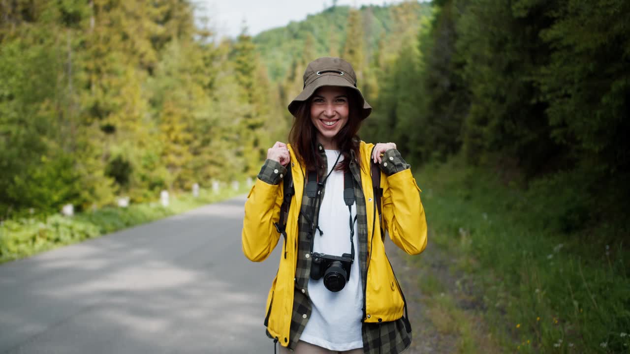 一个穿着特殊徒步旅行服装的旅游女孩的肖像，与穿着黄色夹克的相机一起，她在山间森林的背景下看着相机视频素材