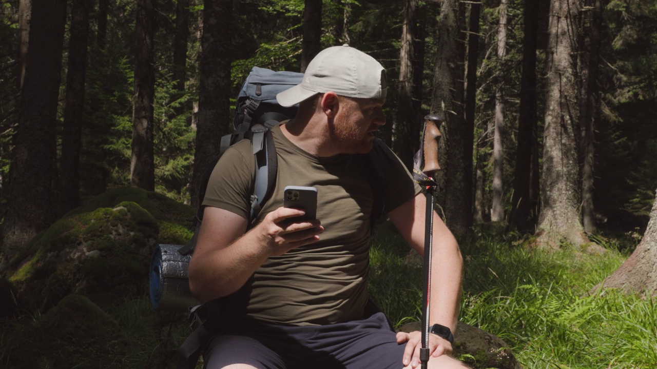 成年男子带着智能手机背包徒步旅行手杖坐在绿色的森林里视频下载