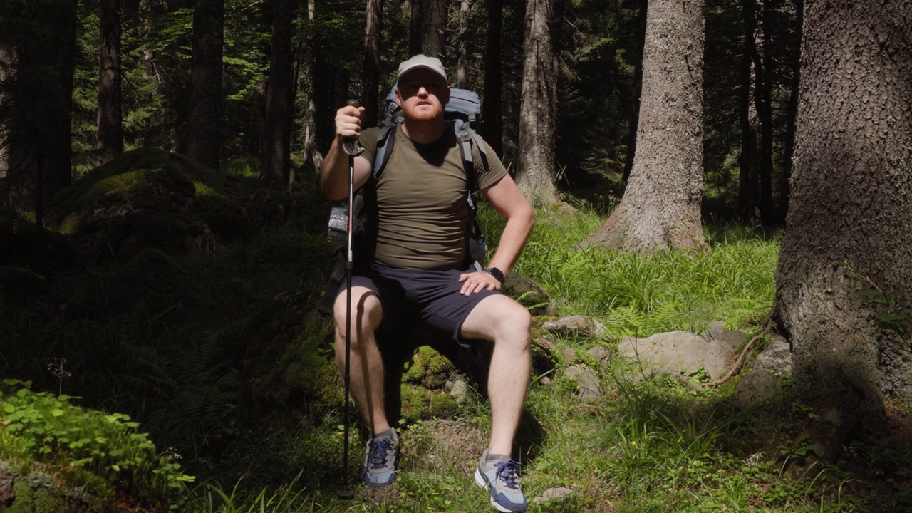 一个成年男子背着背包，把登山杖放在绿色森林的石头上视频素材