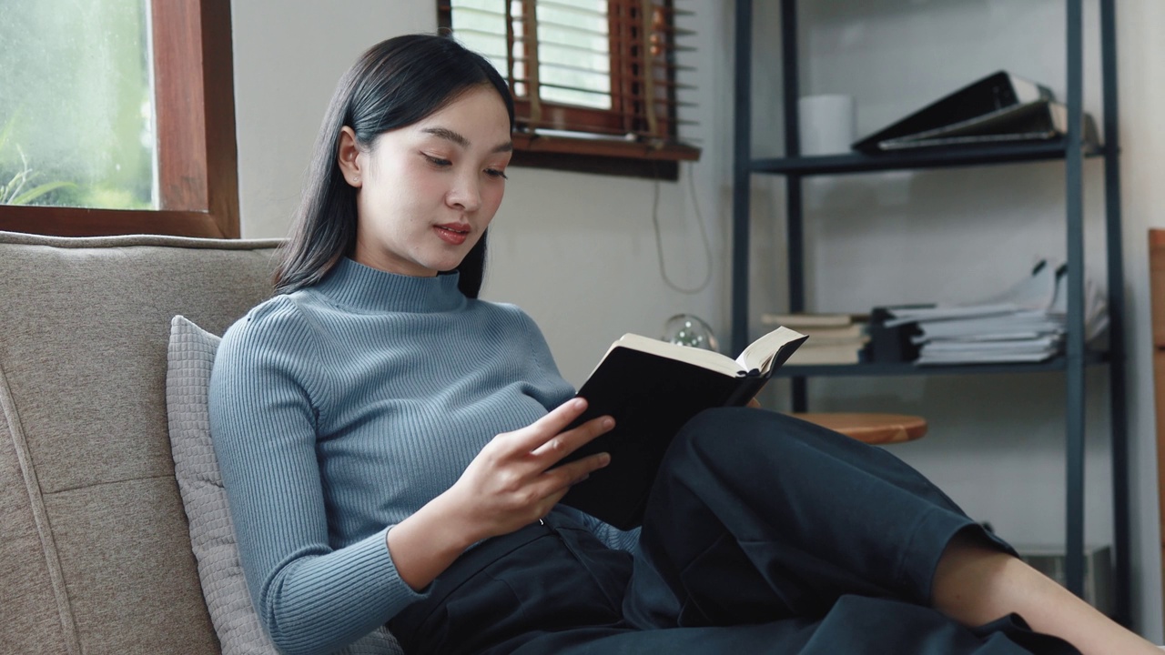 一个亚洲女人正坐在家里的客厅里看书。她在周末度过她的空闲时间。她是公司的雇员。长假期。长假假期。视频素材