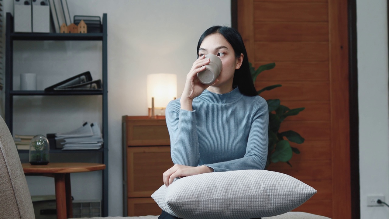 一位亚洲妇女正坐在家里客厅的沙发上喝水。她把业余时间都用在周末休息上。她是公司的雇员。长假期。长周末。视频素材