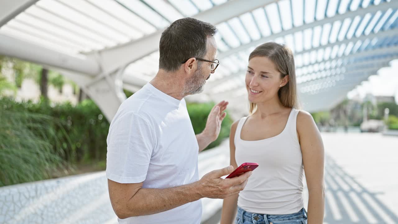 自信的父亲和可爱的女儿，挥舞着智能手机，在充满绿色的可爱公园里享受着酷炫的自拍，他们幸福的微笑反映了他们对家庭纽带的深厚爱。视频素材