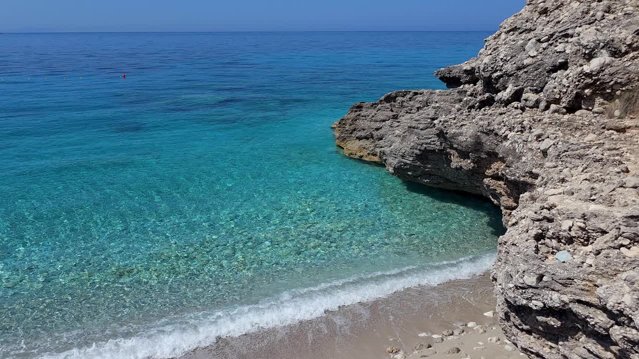 在平静的海滩上闪亮的一天，蔚蓝的海水轻轻地洗过岩石，在阿尔巴尼亚创造了一个宁静的海岸撤退视频素材