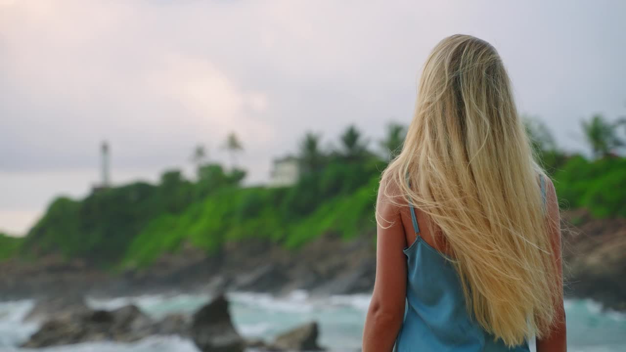 穿蓝色连衣裙的金发女郎在沙滩上漫步。女人欣赏海景，灯塔，在海上找到宁静。健康，心理健康通过大自然的美丽。旅游，休闲，热带环境的生态疗法。视频素材