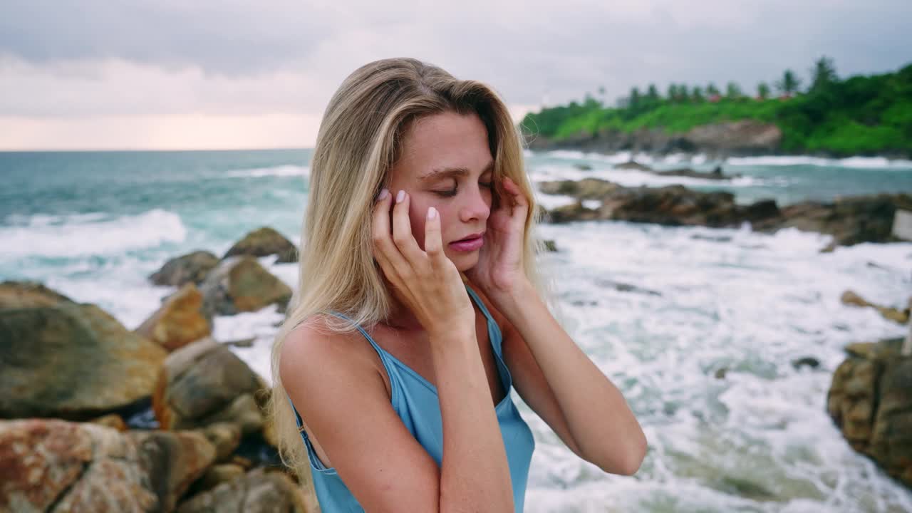 女人在岩石海岸中沉思。温柔的海浪背照阳光，宁静的时刻，自我关怀。金发碧眼，海风吹拂，黎明时分宁静的孤独。年轻的女士感受风，与自然联系视频素材