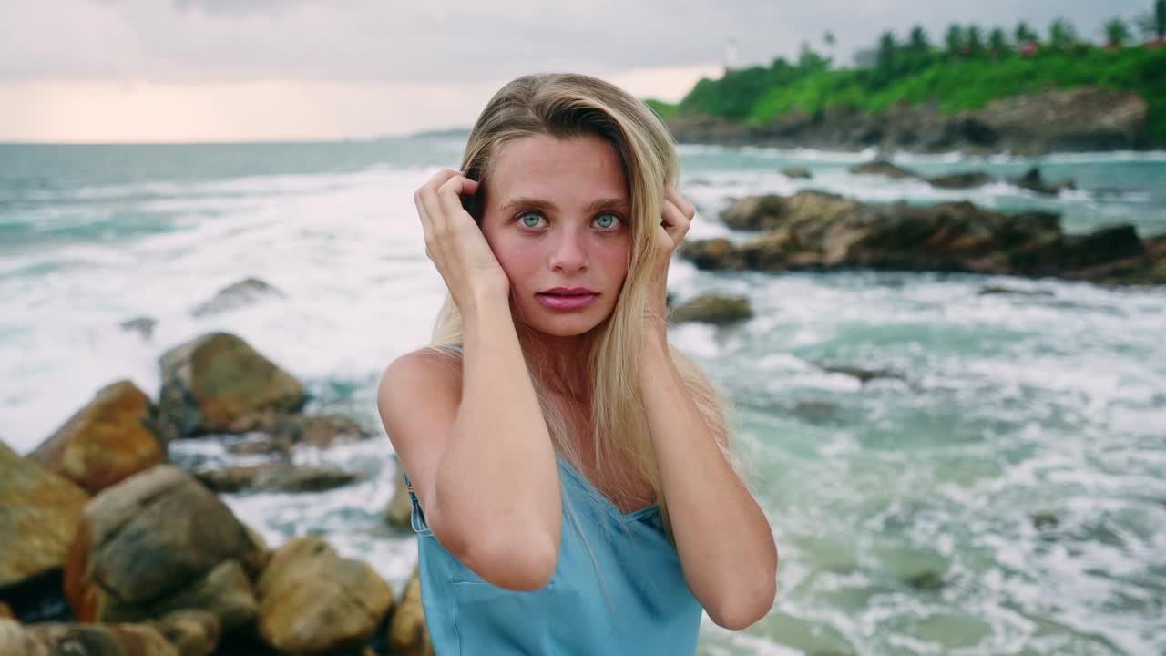 身穿蓝色连衣裙的女人站在悬崖上，海浪拍打着下面的岩石。宁静的女性享受大自然，海风轻轻地吹着头发。宁静，沉思的心情在海边，孤独的时刻。视频素材