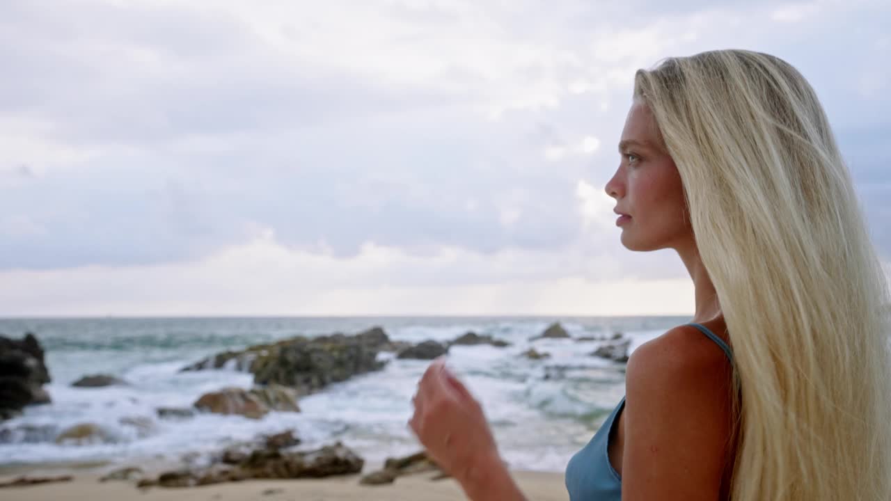 穿着蓝色泳装的金发女子安详地站在岩石嶙峋的海边，海浪汹涌。宁静的海滩景色，反思心情，放松身心。宁静的度假胜地，独自旅行者享受日落。视频素材