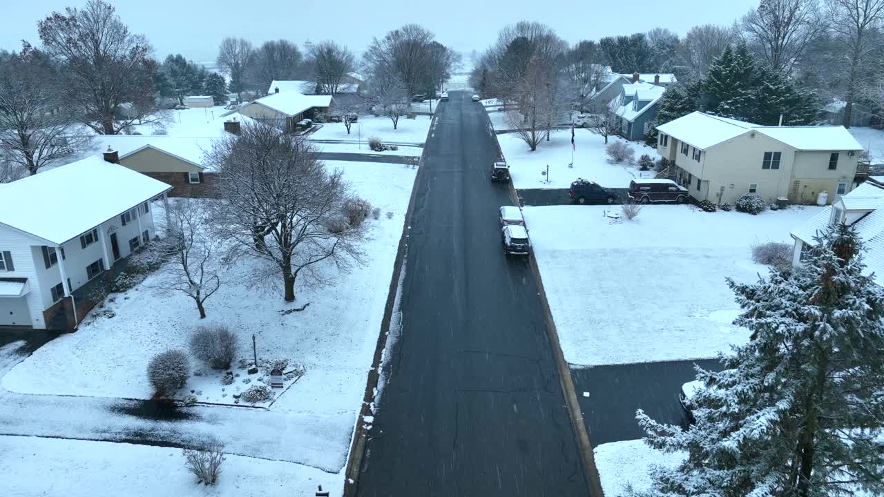 一条白雪皑皑的住宅区街道，两旁是复式住宅和光秃秃的树木。空中飞行在美国附近的小雪。视频素材